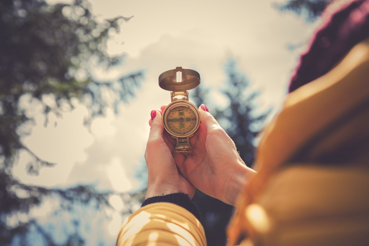 Close-up of hands holding a brass compass outdoors, pointing toward the sky, with pine trees softly blurred in the background — evoking guidance, direction, and intentional navigation.