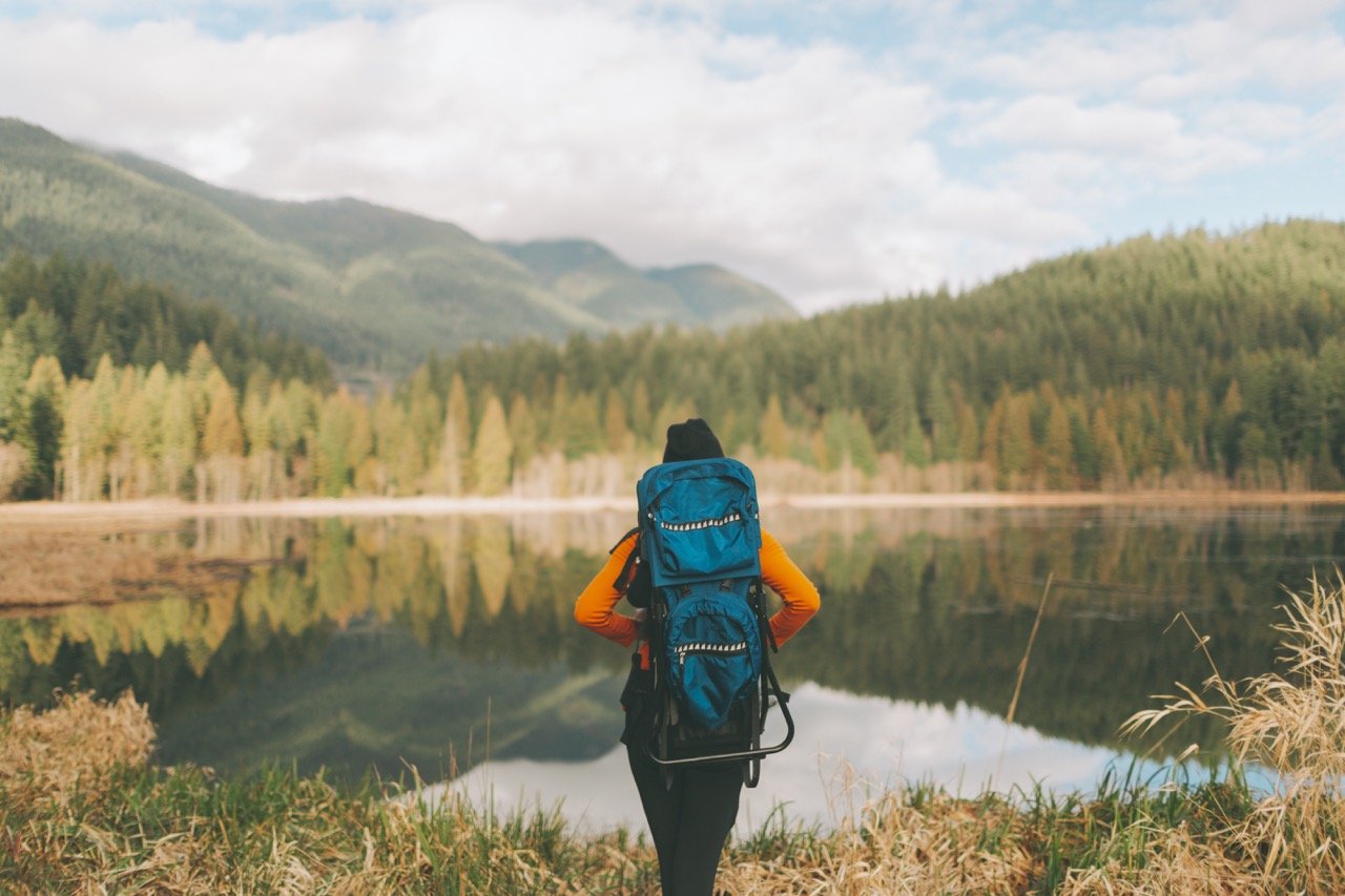 Person wearing an orange jacket and carrying a large blue backpack, standing at the edge of a calm lake, looking out at forested mountains under a cloudy sky.