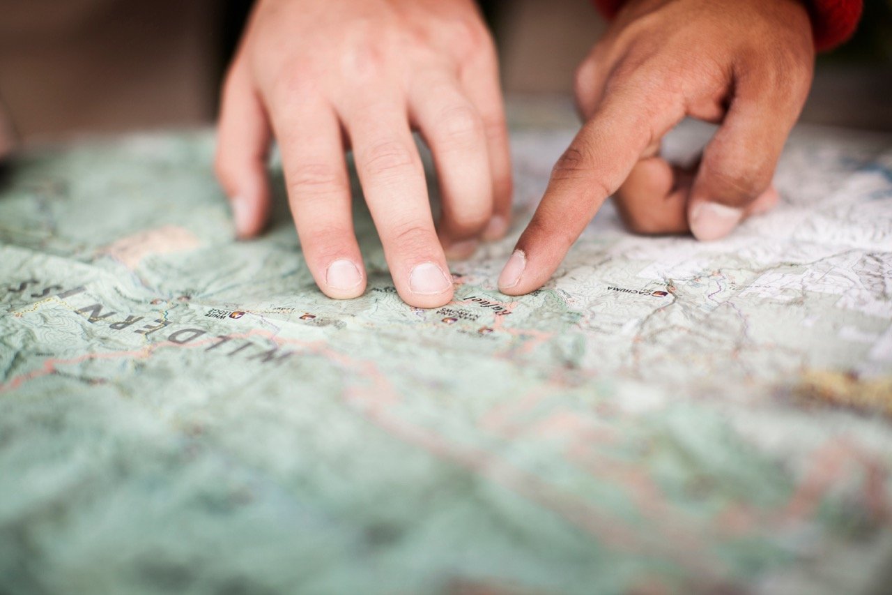 Two pairs of hands pointing to locations on a detailed topographic map, with fingers tracing routes and landmarks — representing collaborative planning, route-finding, and shared strategy.